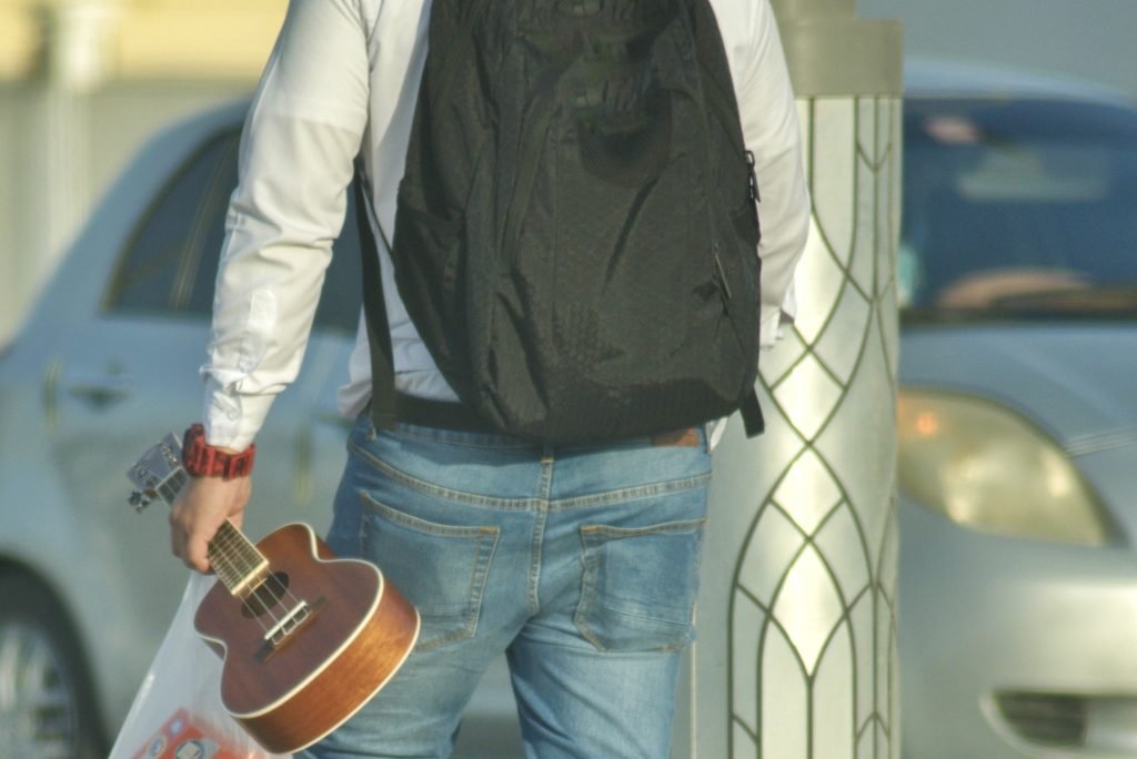 Unrecognizable man from behind with backpack carrying a plastic bag and ukelele on the street